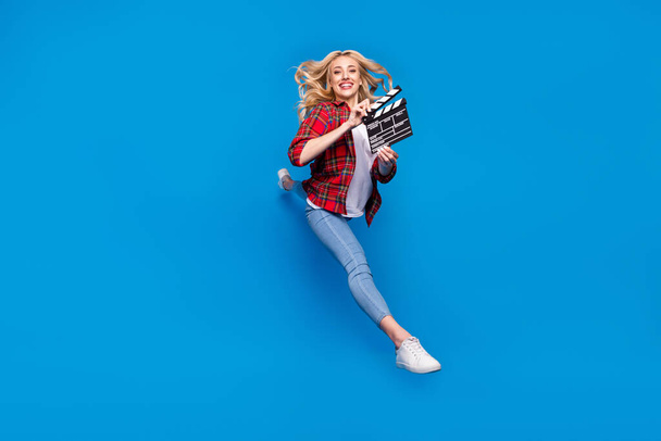Photo of funky sweet young lady wear plaid shirt smiling humping high holding clapperboard isolated blue color background - Foto, Bild