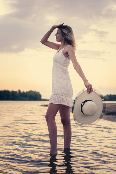 Photo side view of woman with straw hat in her hand. A beautiful young blonde in summer white dress poses against the background of the river bank and holds a hat decorated with a ribbon and flowers - Zdjęcie, obraz