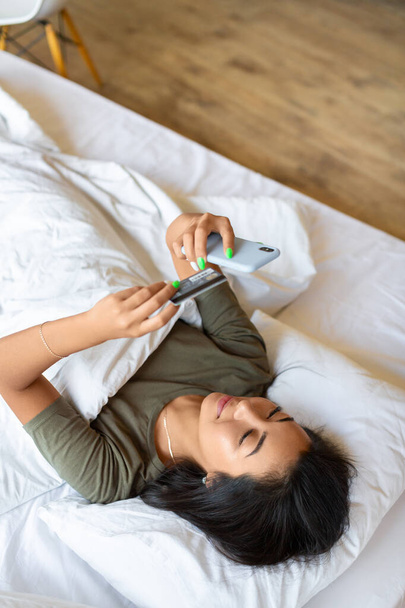Aasialainen nainen tekee verkko-ostoksia nukkuessaan sängyssä. Hän pitää älypuhelinta käsissään ja hyvittää kortin ja valitsee kortin numeron verkkokaupasta. Pystykuva. - Valokuva, kuva
