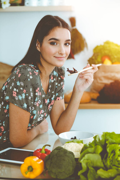 An attractive young dark-haired woman tastes a new recipe for a delicious salad mix while sitting at the table in sunny kitchen. - 写真・画像