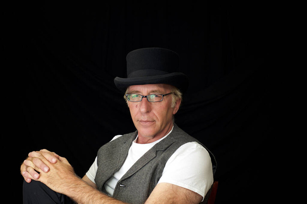 Retrato de estúdio em homem caucasiano usando chapéu superior, colete e óculos, sentado com as mãos no joelho olhando para o espectador. - Foto, Imagem