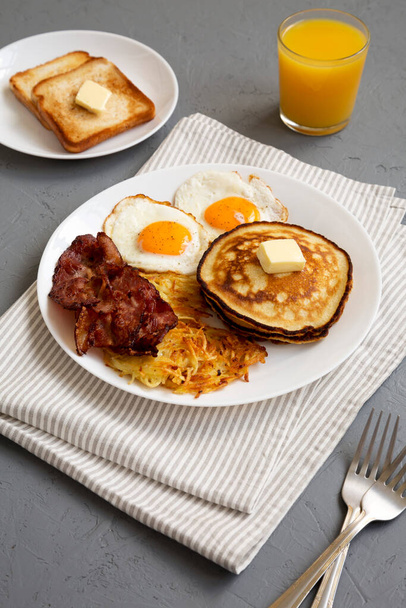 Petit déjeuner américain complet avec bacon, pommes de terre hachées, oeufs et crêpes sur une assiette sur une surface grise, vue à angle bas. - Photo, image