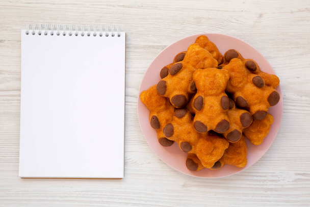 Homemade Sweet Bear Cookies on a pink plate, blank notepad on a white wooden background, top view. From above, flat lay, overhead.  - Photo, Image