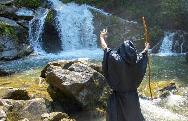 Portret van een bejaarde monnik van 45-50 jaar met een baard in een zwarte soutane met een staf, tegen de achtergrond van een waterval en een bos, selectieve focus. Onderwerp: hekserij en magie, aflopend water uit een steen. - Foto, afbeelding