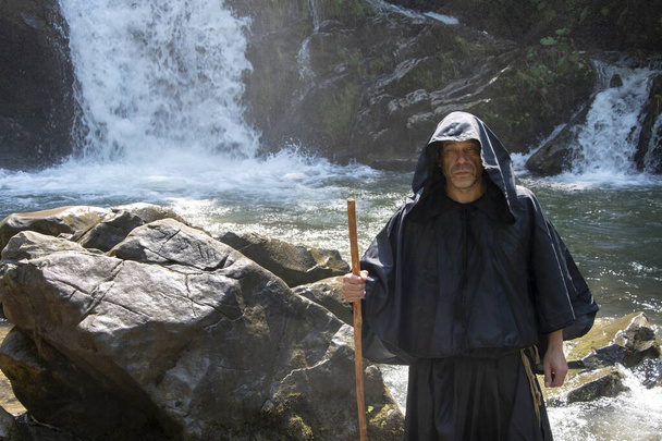 Portrait of an elderly monk 45-50 years old with a beard in a black cassock with a staff, against the background of a mountain waterfall and a forest, selective focus. Concept: witchcraft and magic, descending water from a stone. - Photo, Image