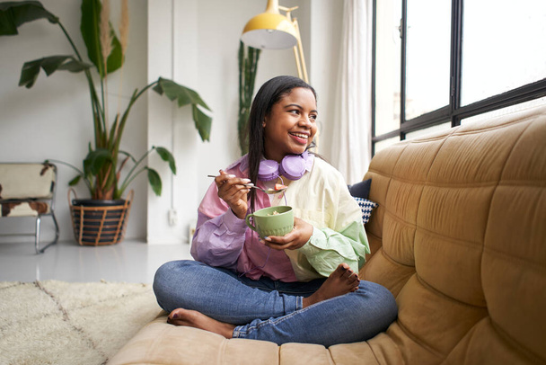 Jeune femme souriante et en bonne santé allongée sur un canapé dans un salon moderne et lumineux, mangeant du lait avec des céréales à l'air détendu et confortable. - Photo, image