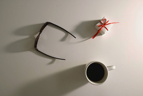 Overhead shot of white cup of coffee next to sunglasses and small gift with red bow on white surface with light shadows, copy space - Fotografie, Obrázek
