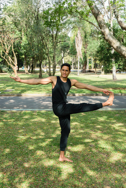 Braziliaanse jongeman die yoga beoefent op een evenwichtige positie in een openbaar park op een zonnige dag. - Foto, afbeelding