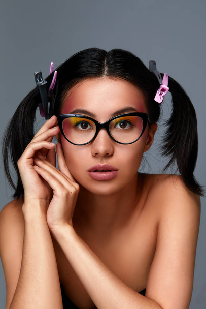 Portret van een Aziatisch meisje in bril, een mooi vrouwelijk gezicht met een schone en gezonde huid, donker haar met vlechtjes, close-up op een grijze achtergrond. - Foto, afbeelding
