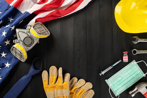 Happy Labor Day during breakout coronavirus. Several constructor work tools, labor COVID-19 vaccine, medical face mask protective and American flag with copy space on black wood background - Foto, Bild
