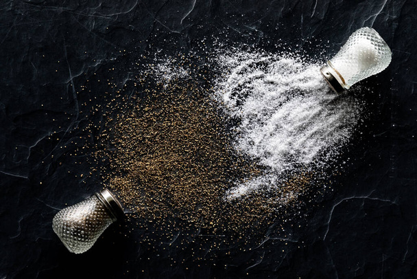Top down view of salt and pepper shakers tipped over with salt and pepper spilled out onto black slate. - Photo, Image
