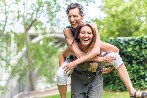 Mature woman giving piggyback to mature friend female in the park - Photo, Image