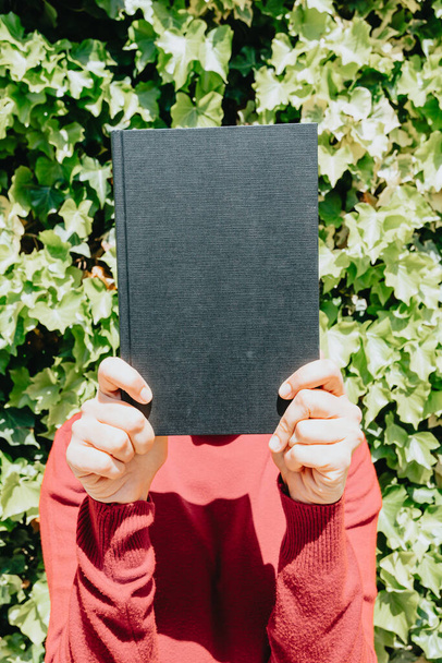 A funny portrait of a girl in a red sweater holding a black book up her face outdoors on a sunny day - Foto, afbeelding