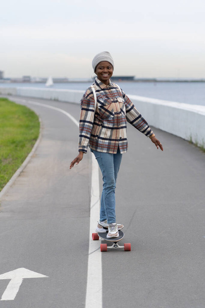 Smiling african girl on skateboard. Happy skateboarder female enjoy longboarding in early spring. - Фото, зображення