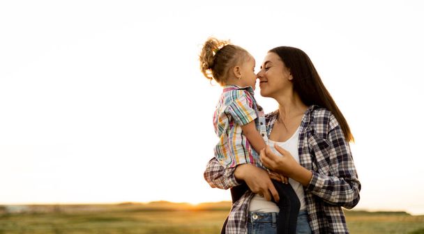 Szczęśliwa matka całuje swoją córeczkę na zielonej łące o zachodzie słońca. Młoda kobieta trzyma swoje dziecko. macierzyństwo i dzieciństwo będą się dobrze bawić w przyrodzie. Dzień Matki. Szczęśliwa koncepcja rodziny. - Zdjęcie, obraz