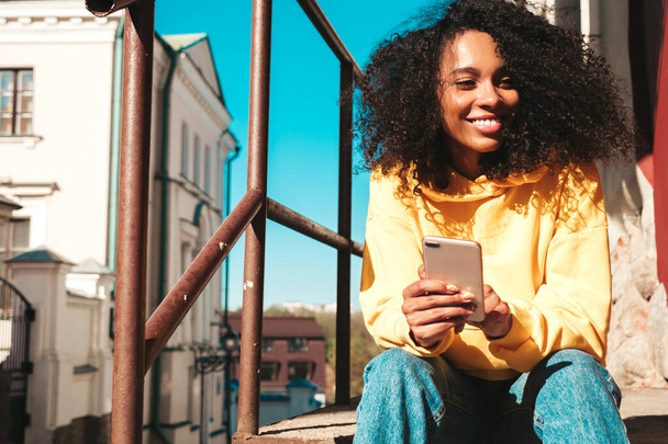 Beautiful black woman with afro curls hairstyle.Smiling model in yellow hoodie. Sexy carefree female posing on the street background. Looking at smartphone screen, using apps - Valokuva, kuva