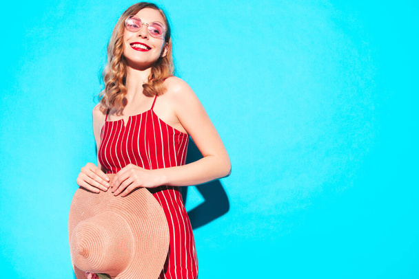Fiatal gyönyörű mosolygós nő divatos nyári piros hipster overálban. Szexi gondtalan nő pózol a kék fal mellett a stúdióban. Pozitív modell szórakozás napszemüvegben és kalapban - Fotó, kép