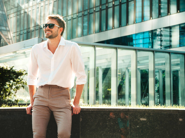 Portrait of handsome smiling stylish hipster lambersexual model.Modern man dressed in white shirt. Fashion male posing in the street background near skyscrapers in sunglasses - Fotoğraf, Görsel
