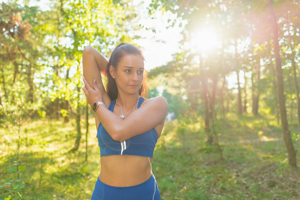 Woman runner stretching arms before exercising in a summer park morning.Young athletic female warming up body before running Caucasian person warm up jogging.Front view. - Foto, afbeelding