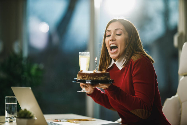 Szczęśliwa młoda kobieta dmucha świece na torcie urodzinowym podczas internetowej imprezy urodzinowej z przyjaciółmi. - Zdjęcie, obraz