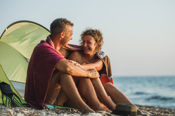 Una joven pareja cariñosa divirtiéndose en la playa y disfrutando de la puesta de sol envuelta en una manta. - Foto, Imagen