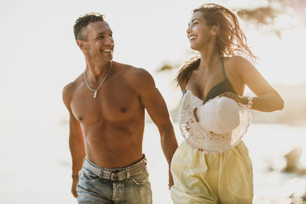 Ευτυχισμένο περιστασιακό ζευγάρι που κρατιέται χέρι-χέρι και τρέχει στην παραλία απολαμβάνοντας καλοκαιρινές διακοπές. - Φωτογραφία, εικόνα