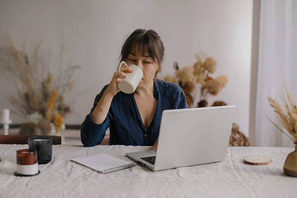 Occupato focalizzato donna asiatica freelance bere tè o caffè mentre si lavora in remoto sul computer portatile da casa - Foto, immagini