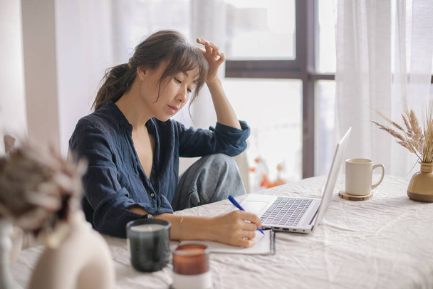 Atraktivní asijská žena dálkově pracuje z domova kanceláře na notebooku, dělat si poznámky v notepadu - Fotografie, Obrázek
