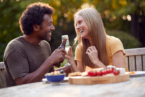 Mature Couple Celebrating With Beer And Champagne As They Sit At Table In Garden With Snacks - Фото, изображение
