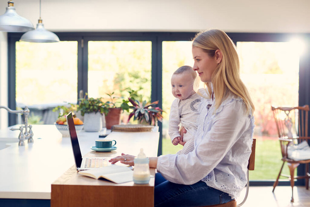 Working Mother Using Laptop At Home Whilst Holding Smiling Baby Son On Knee - Valokuva, kuva