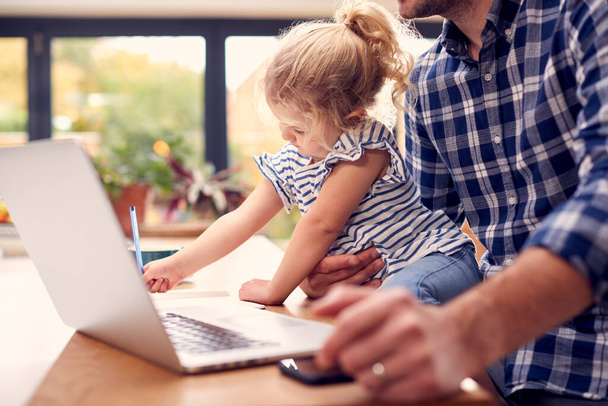 Working Father Using Laptop At Home On Kitchen Counter Whilst Looking After Young Daughter - 写真・画像