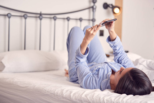 Γυναίκα ξαπλωμένη στο κρεβάτι με κινητό τηλέφωνο φορώντας πιτζάμες - Φωτογραφία, εικόνα