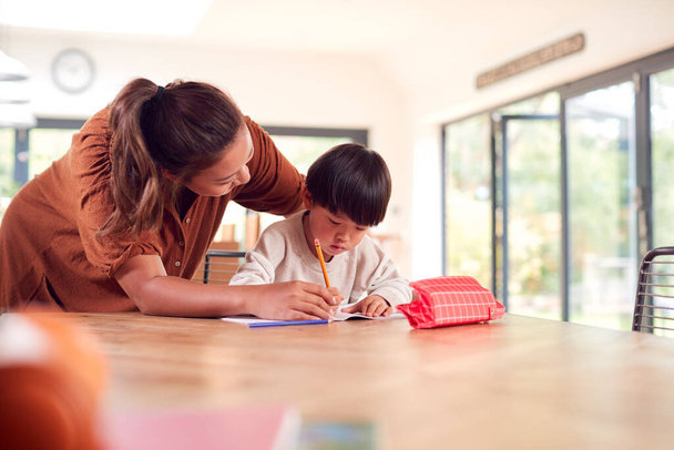 Asian Mother Helping Home Schooling Son Working At Table In Kitchen Writing In Book - 写真・画像