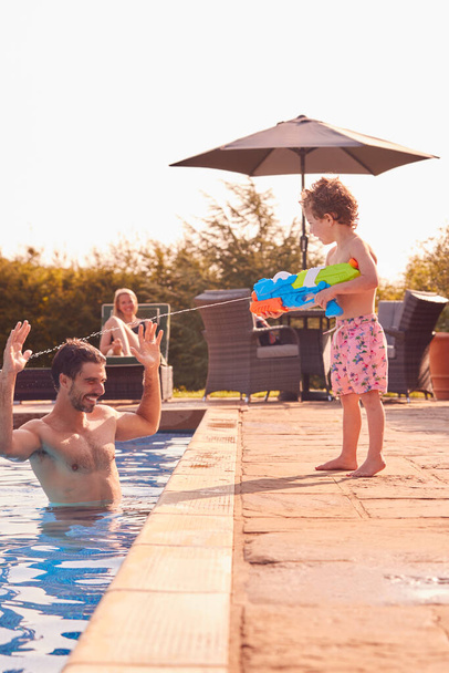 Son Squirting Father With Water Pistol Playing In Swimming Pool On Summer Vacation - Foto, imagen