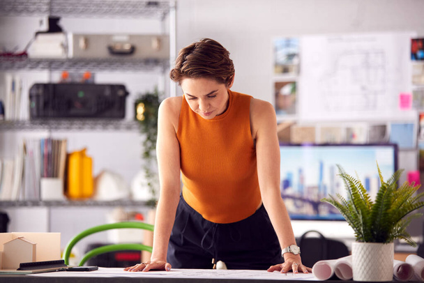 Portrait Of Female Architect Standing At Desk In Office Looking At Plans Or Blueprints - Foto, imagen