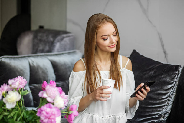 Young redhead woman using mobile phone while sitting on velvet sofa at home. Focused beautiful european girl hold paper cup. Interior of modern apartment with bouquet of peony flowers - Photo, image