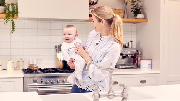 Mother At Home In Kitchen Holding Smiling Baby Son - Fotografie, Obrázek