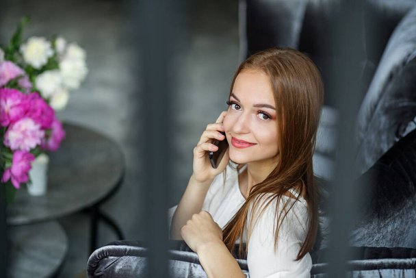 Jeune femme rousse parlant sur un téléphone portable assis sur un canapé en velours à la maison. Souriant belle fille européenne regardant la caméra. Intérieur de l'appartement moderne avec des fleurs de pivoine - Photo, image