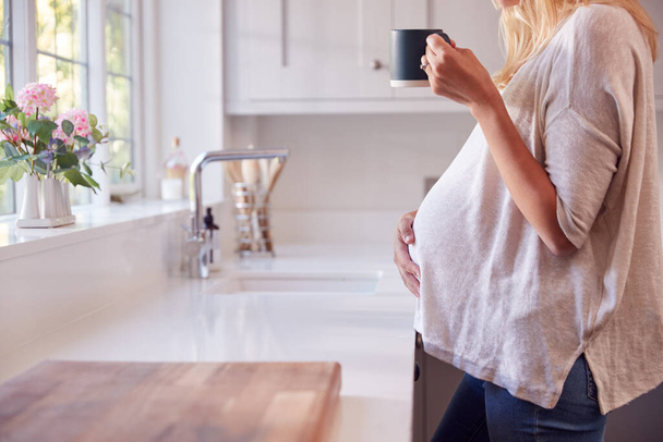 Close Up Of Pregnant Woman Standing By Kitchen Window Touching Stomach Relaxing With Hot Drink - Foto, afbeelding