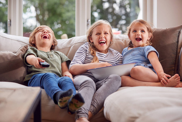 Three Children Sitting On Sofa At Home Laughing And Watching TV With Popcorn - 写真・画像