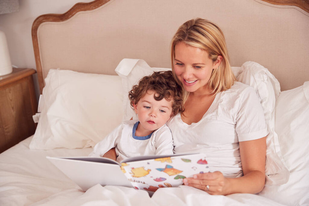Mother In Bedroom Looking At Picture Book With Young Son Wearing Pyjamas - Foto, Imagem
