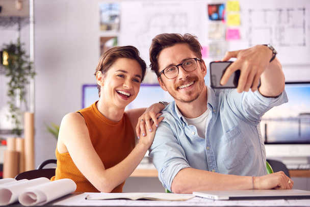 Male And Female Architects In Office Sitting At Desk Posing For Selfie Together - Foto, imagen