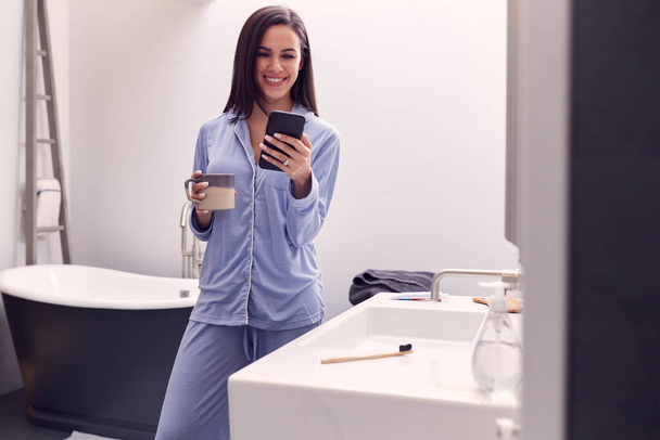 Woman Wearing Pyjamas With Hot Drink At Home Checking Mobile Phone In Modern Bathroom - Photo, Image