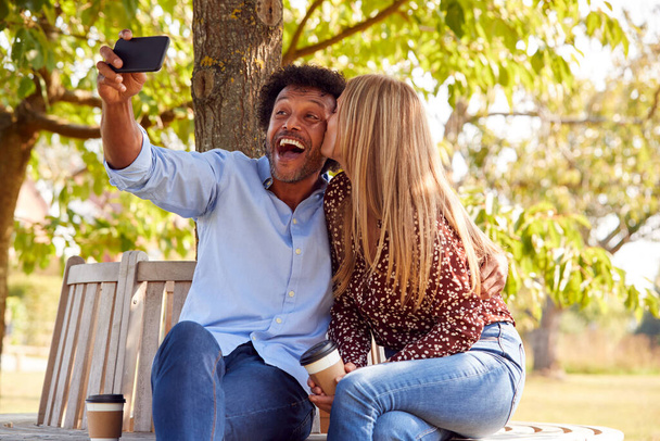 Kissing Mature Couple Posing For Selfie On Mobile Phone Sitting On Seat In Park - Foto, afbeelding