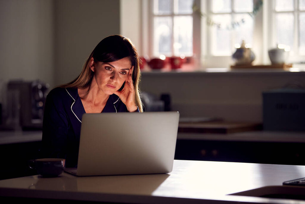 Unhappy Woman Wearing Pyjamas Sitting In Kitchen At Night Using Laptop - Foto, Imagem