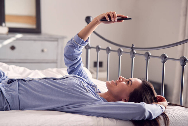 Woman Wearing Pyjamas Taking Selfie On Mobile Phone Lying On Bed - Foto, imagen