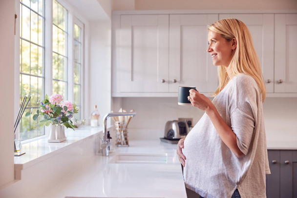Pregnant Woman Standing By Kitchen Window Touching Stomach Relaxing With Hot Drink - Foto, afbeelding