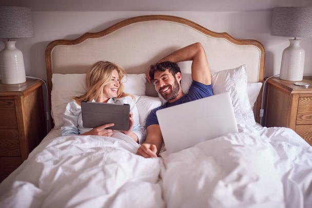 Пара лежащих в постели утром глядя на цифровой планшет и ноутбук - Фото, изображение
