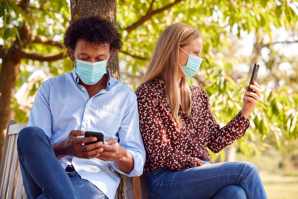 Couple Wearing Masks Meeting In Outdoor Park During Health Pandemic Looking At Mobile Phones - Foto, Imagem