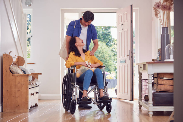 Asian Man Pushing Wife In Wheelchair At Home Back From Shopping Trip With Bag - Foto, imagen